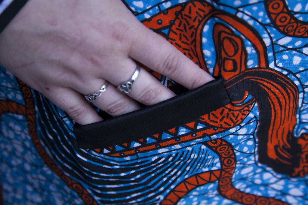 Woman's bomber jacket with African Print fabric, showing a welt pocket. Whimsical pattern with abstracted soda cans surrounded by white splotches on light blue contrasted with orange.