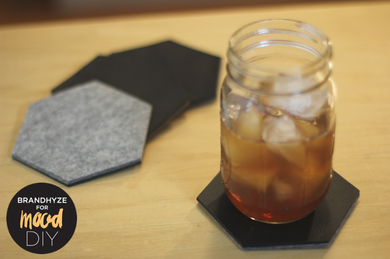 Four reversible leather and felt coasters displayed, each shaped as a hexagon. One side features black leather, while the other is gray felt. An iced beverage in a mason jar sits on one coaster, demonstrating its practicality for iced drinks on a summer day.
