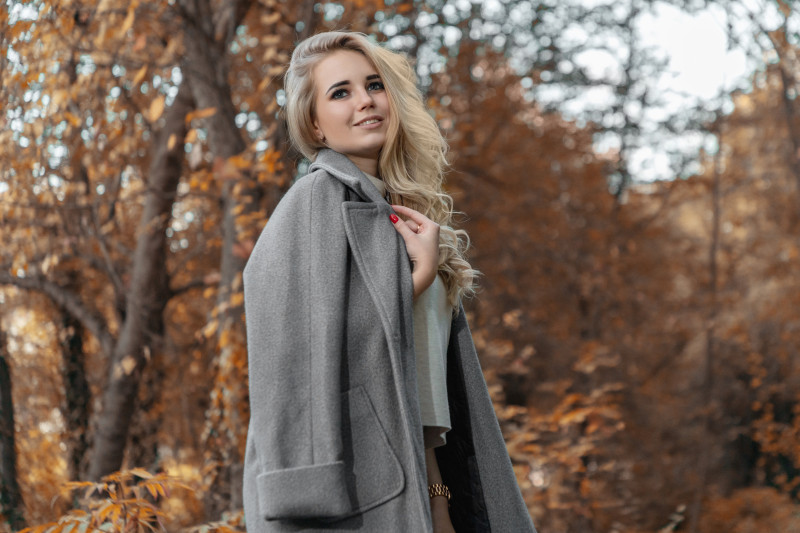 woman in a long coat standing outside by trees covered in fall leaves