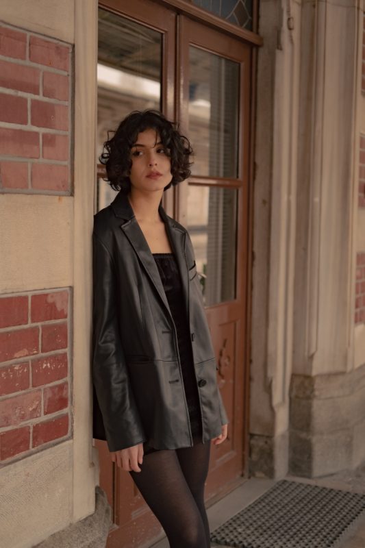 woman posed against door outside wearing a black leather blazer and mini dress with sheer tights