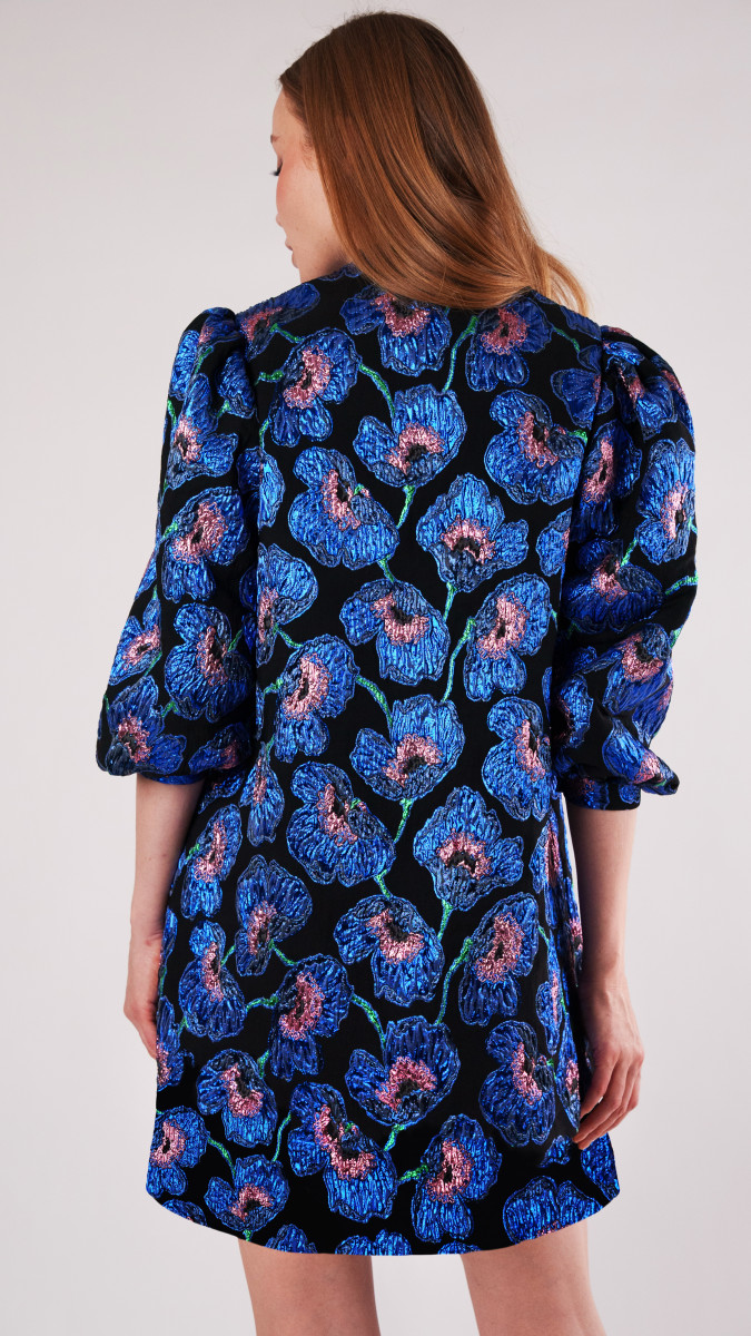 Model posed for a back view of emberly mini dress garment with hip slightly arched to the left, arms hanging straight at sides and head looking down at the model's left side. Model wearing brilliant blue brocade fashion fabric featuring poppy floral pattern woven with metallic threads.