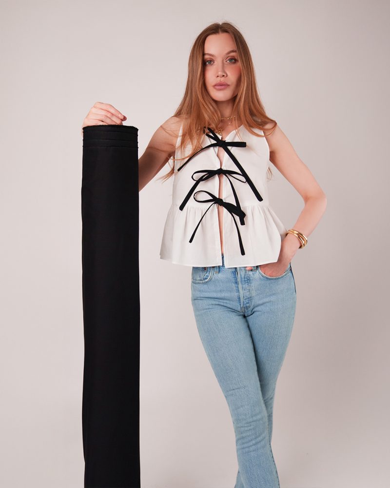 Front view of female model posing by cotton voile fabric roll with one hand in her pocket wearing tie front top and jeans