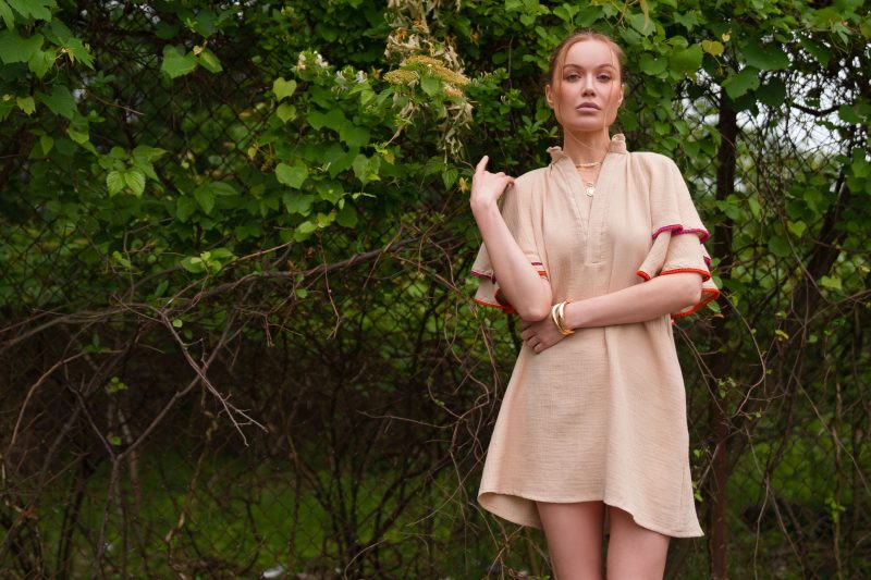 Front view of female model posing outside with greenery wearing a tan colored A-line dress made from double cotton gauze and miniature pom pom trim