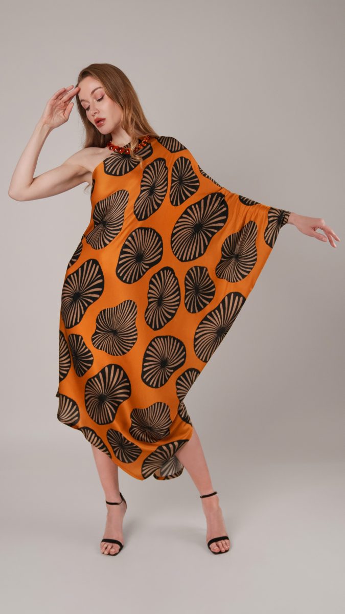 Model posing with one hand on her face wearing orange print open shoulder dress with medallions made from viscose dobby