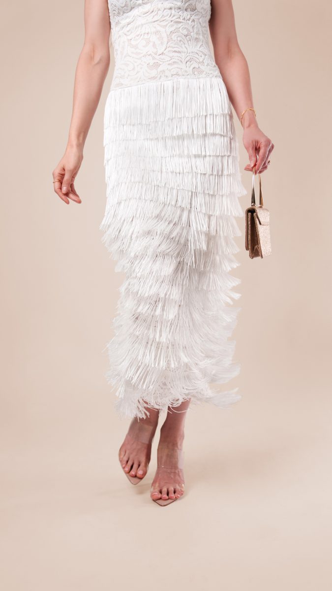 Close-up of Lumiere Strapless Dress showing tiered white fringe details in motion, paired with a chic gold handbag and clear strap heels.