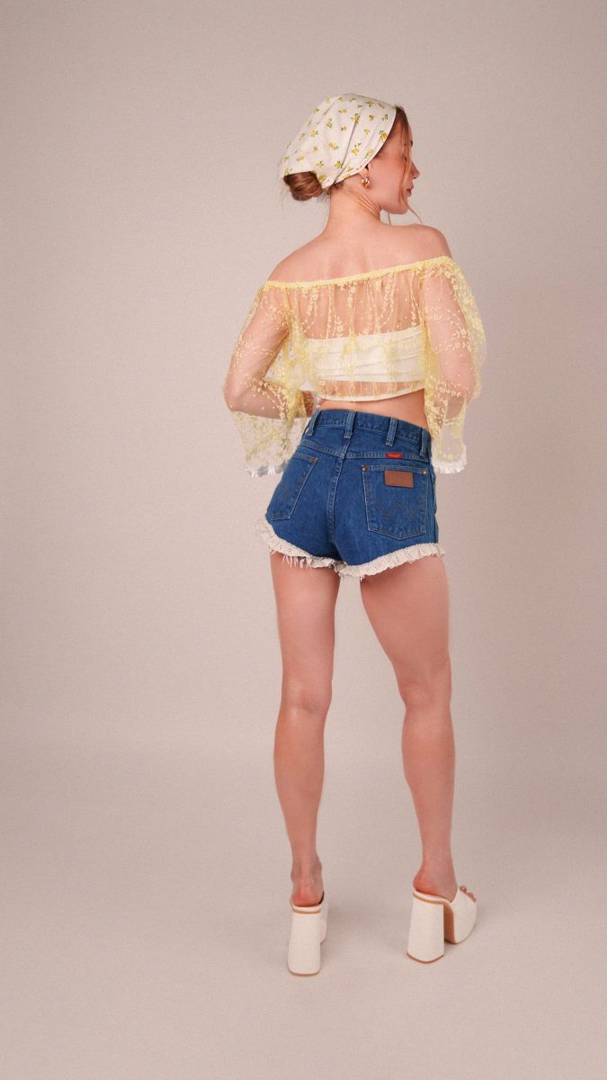Back view of the model highlighting the sheer detail and flared sleeves of the Juno Crop Blouse, sewn from Rit-dyed white floral lace by Mood Fabrics and styled with ruffled denim shorts.