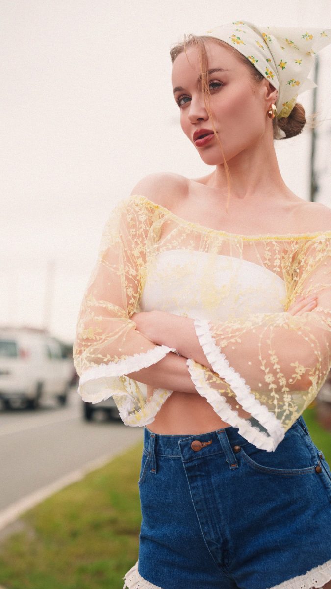 Side view of the model showcasing the off-the-shoulder Juno Crop Blouse made from Rit-dyed white lace floral fabric from Mood Fabrics, paired with distressed denim shorts and gold hoop earrings.
