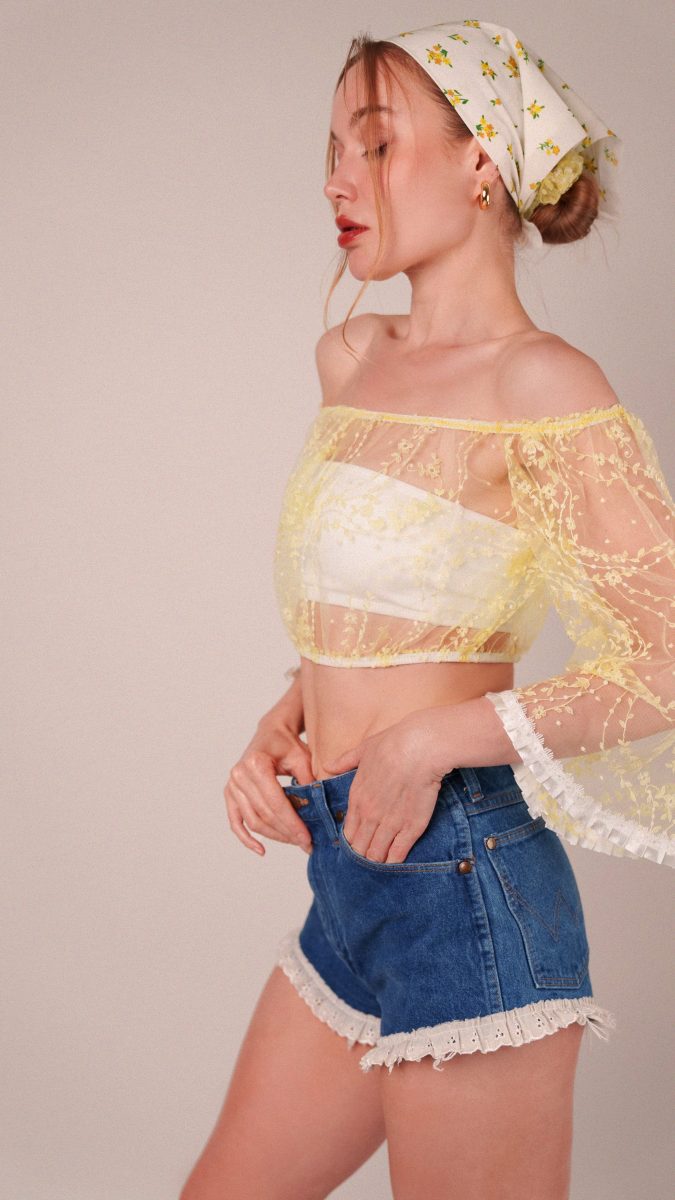 Studio portrait of the model in profile wearing the off-the-shoulder Juno Crop Blouse created with Mood Fabrics’ white lace floral fabric, hand-dyed with Rit Dye for a soft yellow hue.