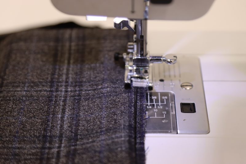 How To Place Hem Under Preser Foot - Super 130s Wool Twill Suiting Made in Italy Charcoal Gray Navy Plaid Deadstock - Mood Fabrics - Oxford Pleated Skirt and Tie Free Sewing Pattern Tutorial - MDF460 - Mood Fabrics Sewciety Blog - Place the hem under the presser foot so that the straight stitch catches only the wrong side of the raw hem and the blind stitches catch the wrong side of the folded edge of the hem.