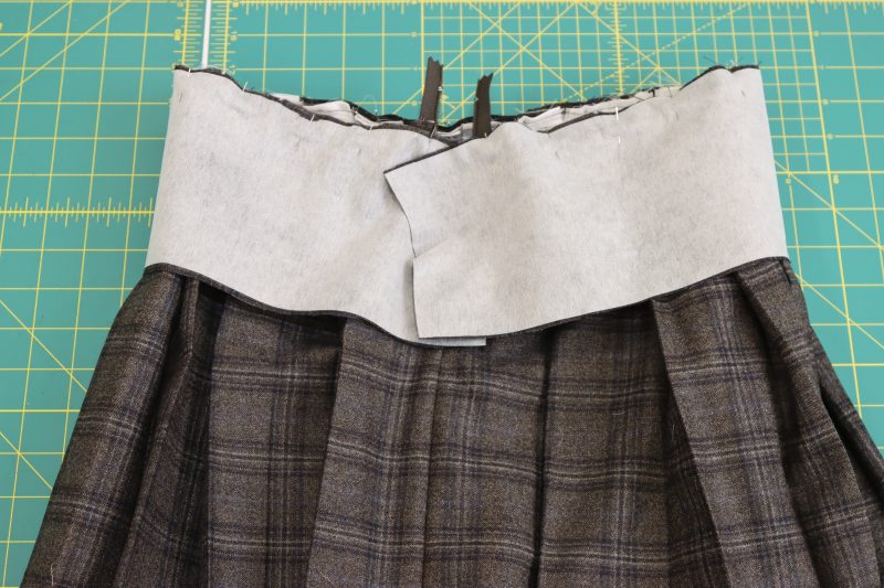 How To Pin Sew Waistband To Raw Edge Of Skirt - Super 130s Wool Twill Suiting Made in Italy Charcoal Gray Navy Plaid Deadstock - Mood Fabrics - Oxford Pleated Skirt and Tie Free Sewing Pattern Tutorial - MDF460 - Mood Fabrics Sewciety Blog - Pin and sew the waistband to the raw edge of the waist opening of the skirt.