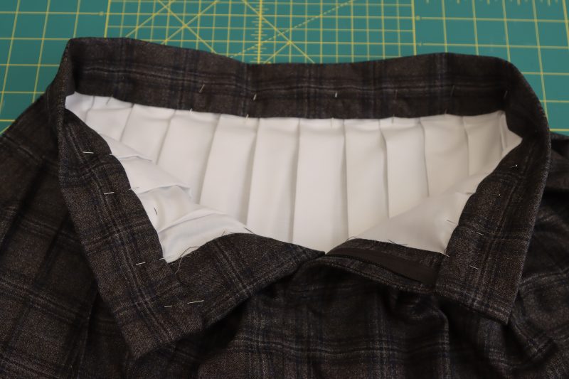 How To Fold Raw Edge Of Waistband Inside - Super 130s Wool Twill Suiting Made in Italy Charcoal Gray Navy Plaid Deadstock - Mood Fabrics - Oxford Pleated Skirt and Tie Free Sewing Pattern Tutorial - MDF460 - Mood Fabrics Sewciety Blog - Fold and pin the inside raw edge of the waistband by 1 inch.