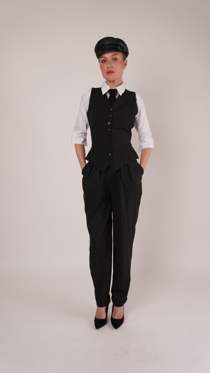 Model wearing the Anne Waistcoat Ensemble styled as a gangster costume, featuring a black pinstripe waistcoat, crisp white shirt, tailored trousers, and black leather cap.