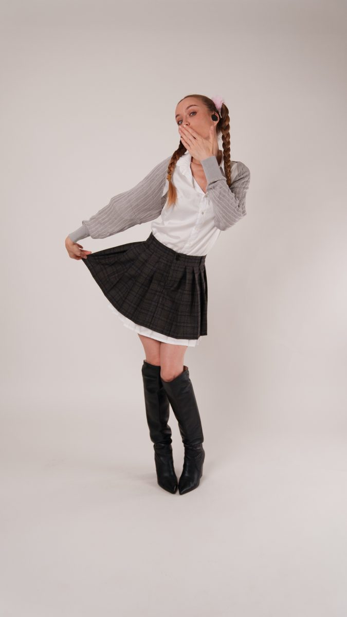 Model in a plaid pleated mini skirt and white button-up shirt with gray sleeves, styled with pink fluffy hair ties in pigtails and knee-high black boots. She poses playfully, holding out the skirt with one hand while covering her mouth in a surprised expression.