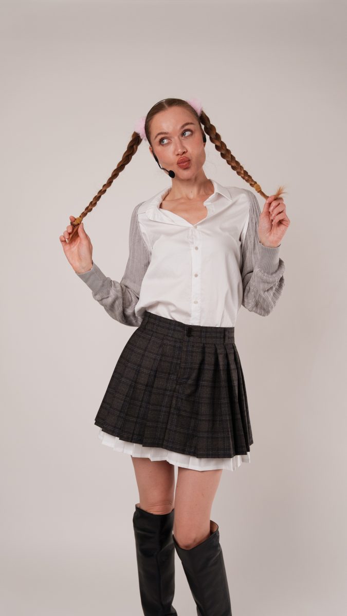 Model wearing a plaid pleated skirt and white button-up shirt with gray sleeves, accessorized with pink fluffy hair ties in braided pigtails. She pulls on her braids while posing with a playful pout, completing the schoolgirl-inspired pop star costume.