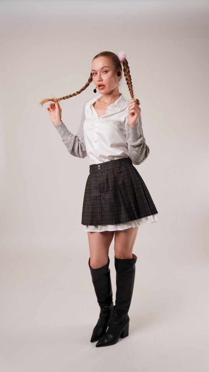 Model styled in the Oxford Skirt free sewing pattern as a Britney Spears schoolgirl Halloween costume. The outfit includes a pleated plaid mini skirt, white button-down shirt, gray sweater sleeves, knee-high black boots, and braided pigtails with pink scrunchies.