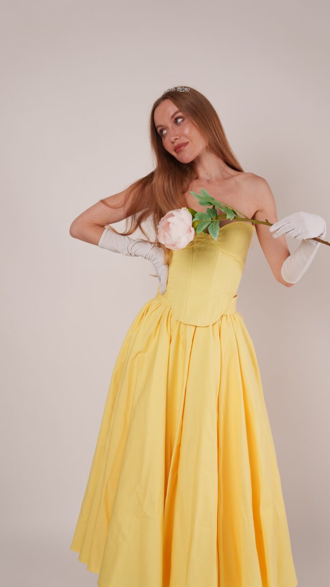 Close-up of the Madison Tea Length Dress in golden yellow with corset-inspired bodice detail. Model holds a rose near her shoulder while wearing a tiara and white gloves, highlighting the fairytale-inspired princess aesthetic.