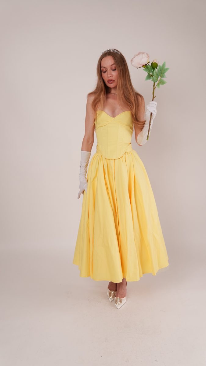Model wearing the Madison Dress free sewing pattern reimagined as a Belle-inspired princess Halloween costume. The strapless yellow gown features a fitted bodice and full skirt, styled with white gloves, a tiara, and a rose prop for a fairytale look.