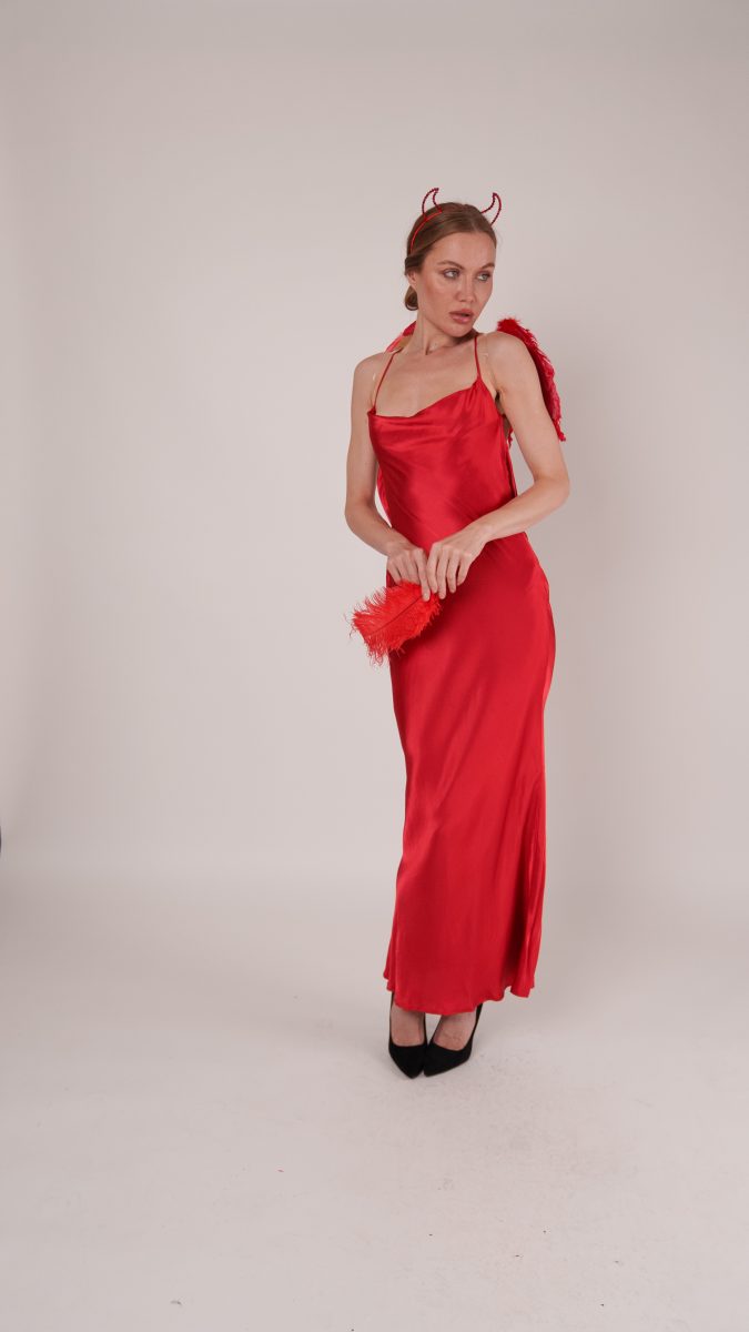 Model wearing a red satin Vixen Slip Dress styled as a devil Halloween costume, accessorized with red horns headband, feathered wings, and black heels while holding a red feather prop.