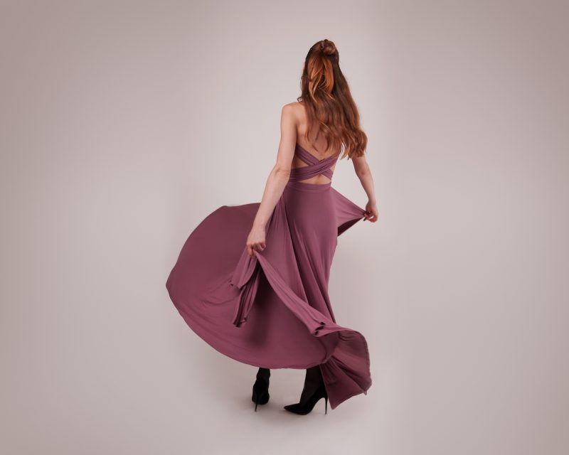 A view from the back of a model wearing the mauve Rhodes Infinity Dress (MDF309), showing the criss-cross convertible straps and the full, sweeping maxi skirt in motion.