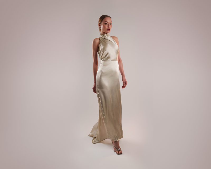 A model wearing the floor-length Ainsley Dress (MDF191), a sleek champagne satin gown with a high cowl halter neckline and a side slit closure detailed with large covered buttons.