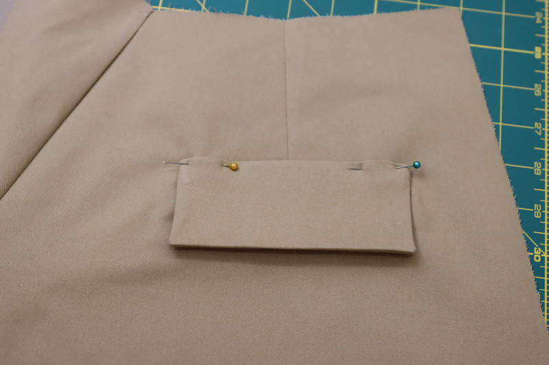 How To Pin Pocket Flaps To Upper Seam Allowance Of Welt Pocket - Stretch Khaki Twill - Mood Fabrics - Lucca Tailored Barrel Leg Pants Free Sewing Pattern Tutorial - MDF524 - Mood Fabrics Sewciety Blog - Pin the pocket flaps to the upper seam allowance of the welt pocket.