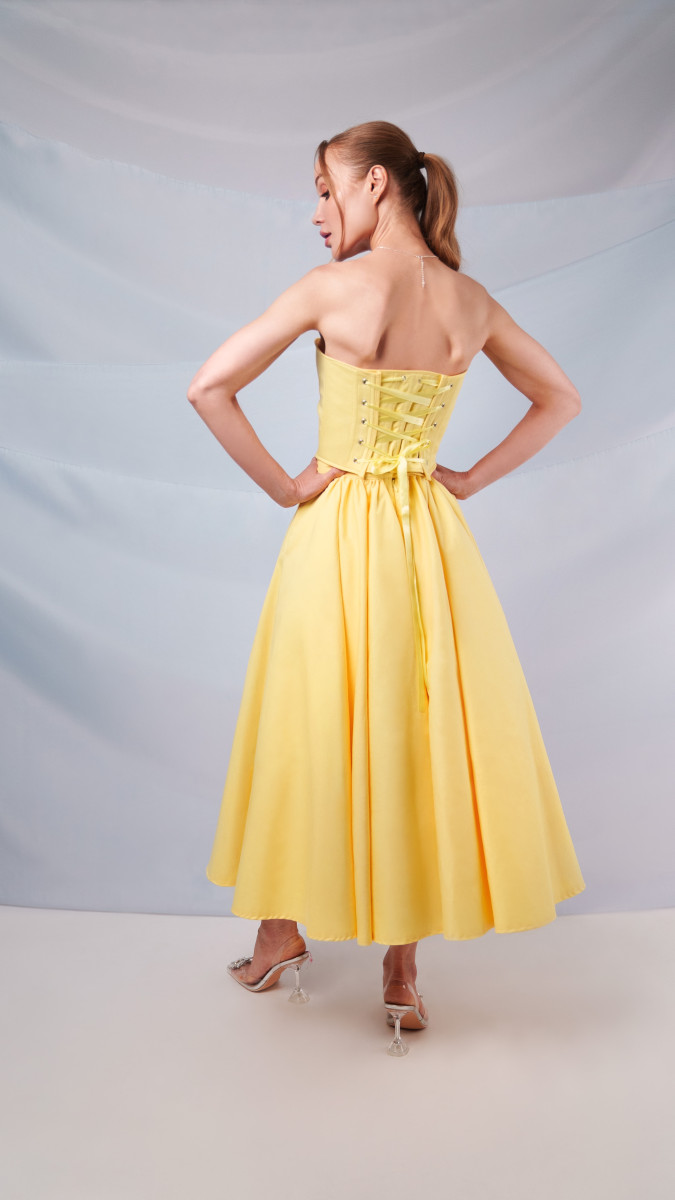 Rear view of the Madison Tea Length Dress in yellow organic cotton twill, showing the traditional lace-up corset back and gathered full skirt.