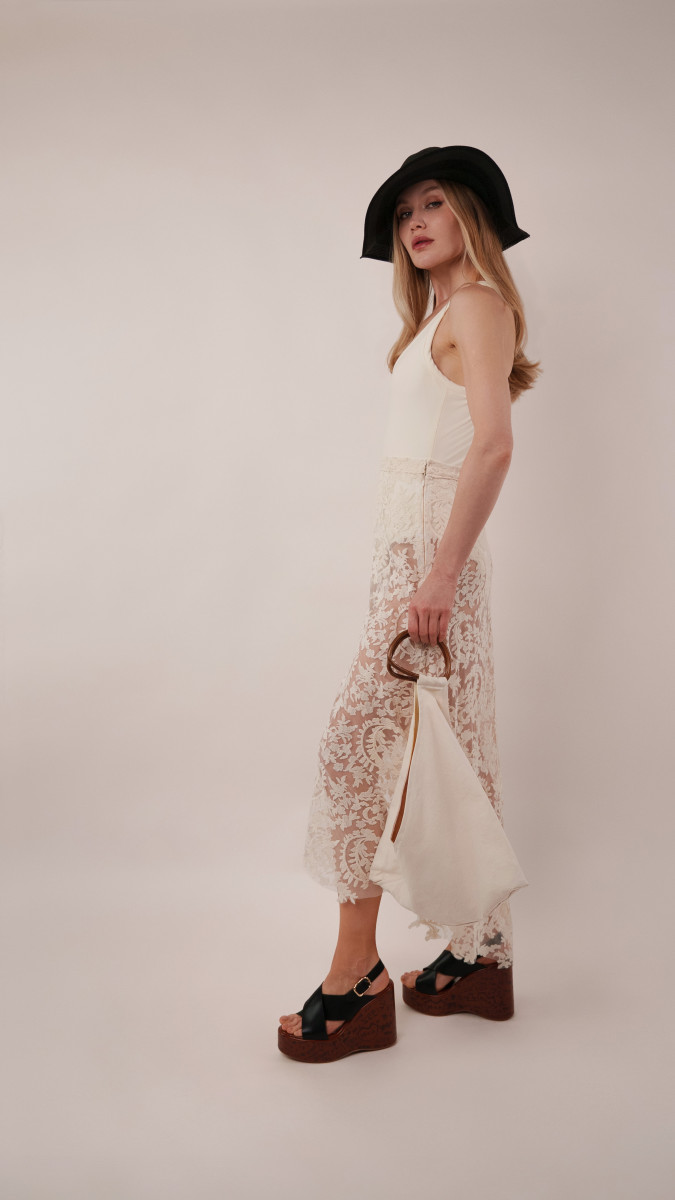 Model in an ivory Clara Bodysuit and high-low lace Taiga Trapeze Skirt with a black sun hat for a sophisticated Vacation Capsule evening outfit.