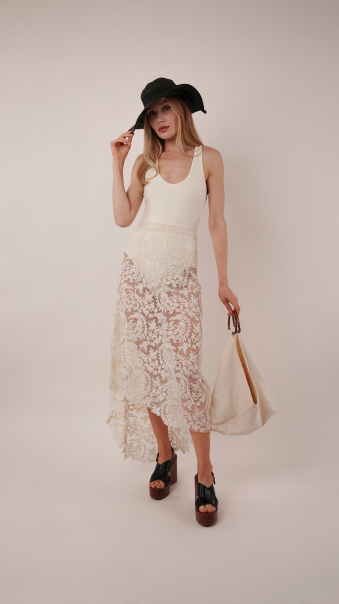 Model in an ivory Clara Bodysuit and high-low lace Taiga Trapeze Skirt with a black sun hat for a sophisticated Vacation Capsule evening outfit.
