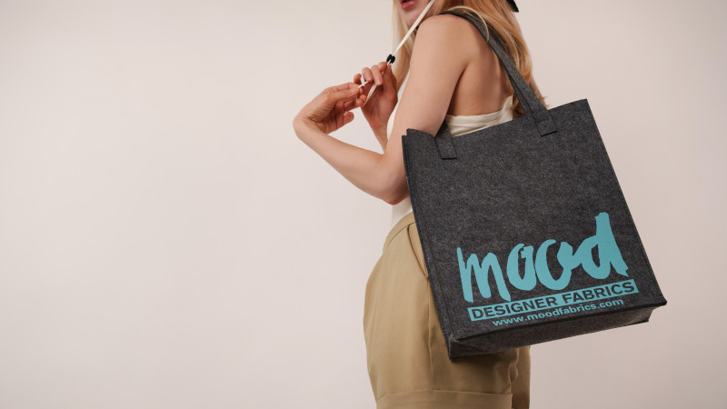 Close-up of a model carrying the Mood Felt Tote Bag in charcoal grey felt with the teal Mood Designer Fabrics logo, a key accessory for the vacation capsule.