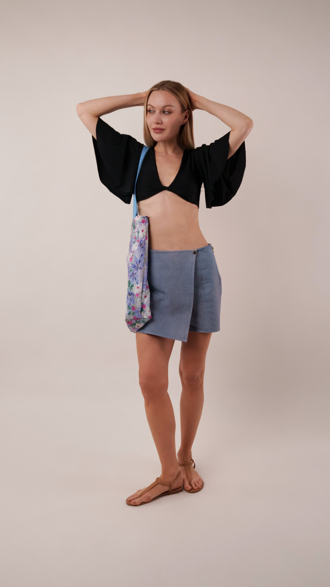 Model wearing a black Reed Swimsuit and blue Biarritz Skort with a floral Spirea Tote Bag for a boardwalk-ready Vacation Capsule look.