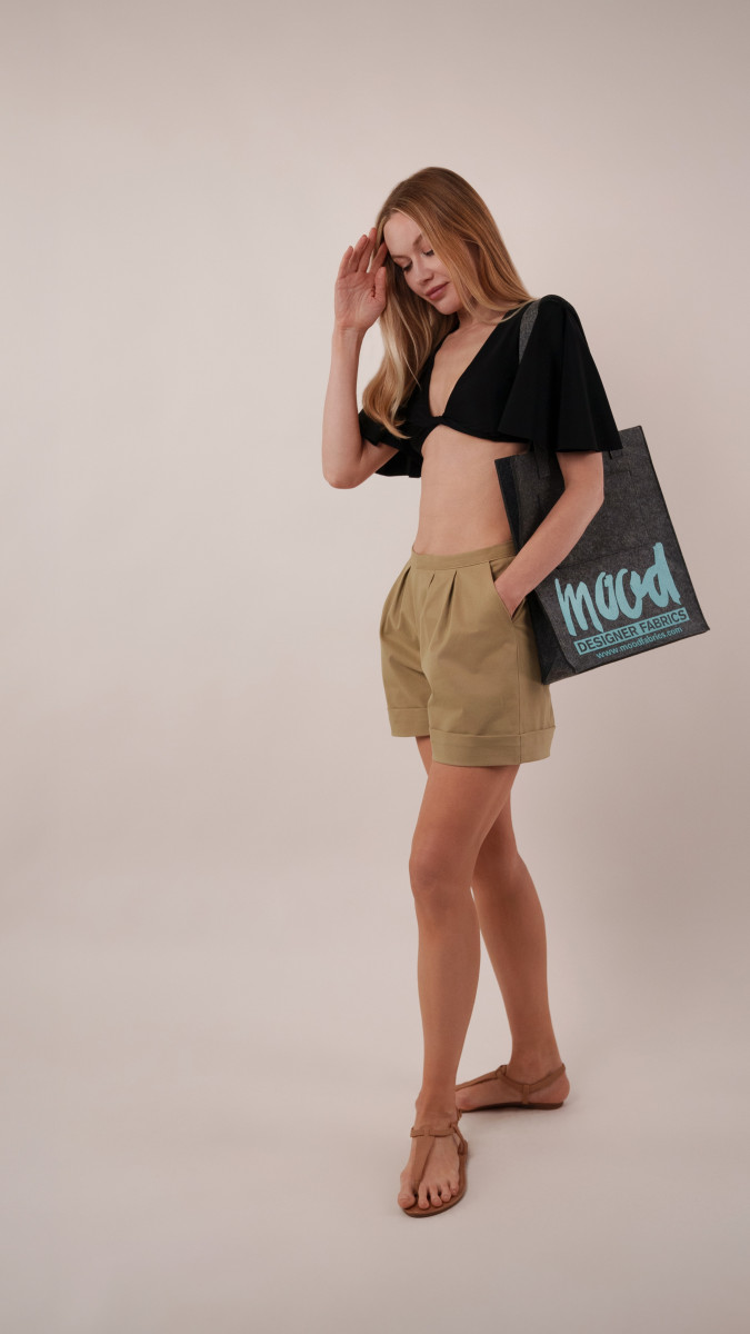 Model wearing a black Reed Swimsuit tucked into khaki Dresden Shorts, styled with a charcoal Spirea Tote Bag for a vacation capsule beach-to-lunch look.