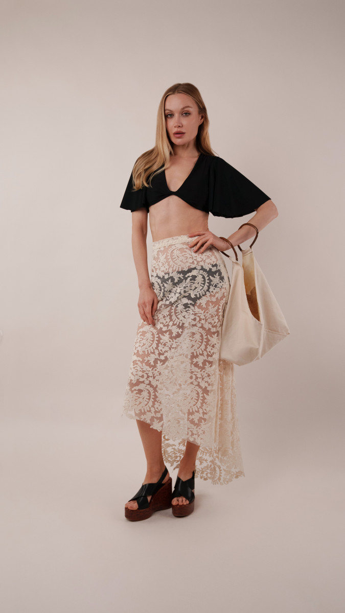 Model wearing the black Reed Swimsuit and high-low lace Taiga Trapeze Skirt with a black sun hat and ivory Spirea Tote Bag for a vacation capsule sunset beach party.