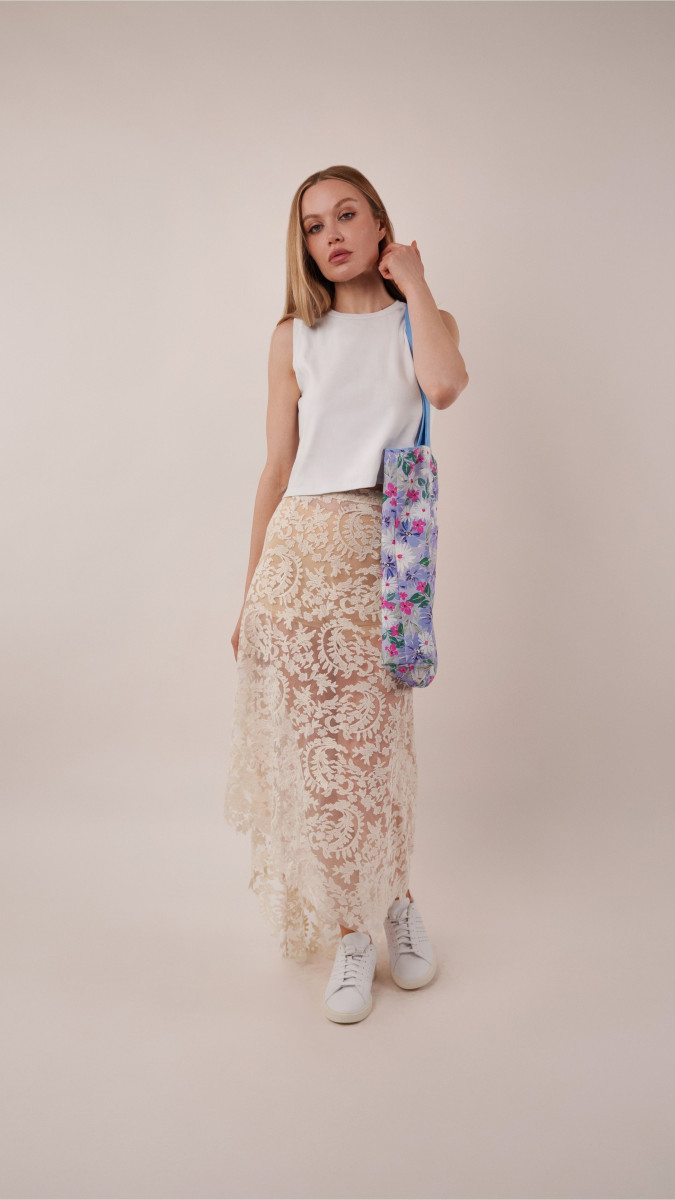 Model in a white Sylvan Tank Top and high-low lace Taiga Trapeze Skirt paired with white sneakers and a floral Spirea Tote Bag for a vacation capsule dinner look.
